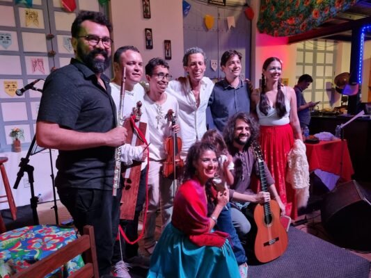 Grupo de músicos posando juntos após uma apresentação, sorrindo para a câmera. Alguns seguram instrumentos, como flauta e violino, enquanto outros têm trajes coloridos. O ambiente é decorado com bandeirolas e elementos de festa típica, em um espaço interno iluminado.