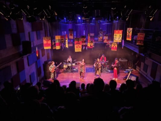 Cena de um show musical em um teatro, com seis músicos no palco iluminado por luzes roxas e amarelas. Bandeiras coloridas decoram o fundo, enquanto a plateia assiste na penumbra.