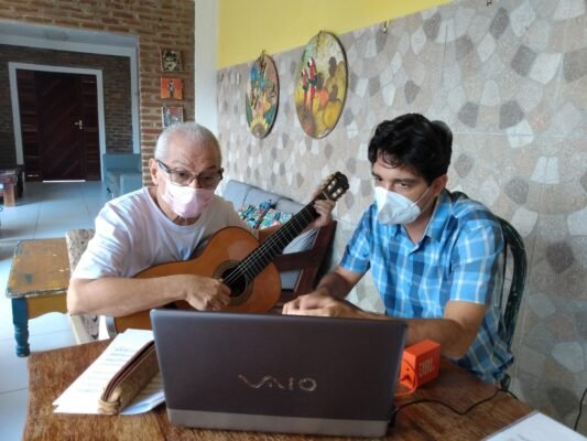 Antonio, com máscara facial, está tocando violão ao lado de Francisco Andrade, que também utiliza máscara. Ambos estão concentrados diante de um laptop sobre uma mesa, acompanhados por partituras musicais. O ambiente tem paredes decoradas por peças artísticas e móveis.