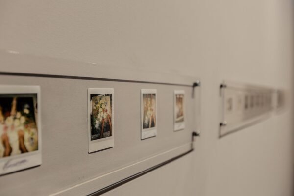 Vista da exposição de D.C. no Instituto Çarê. Uma série de fotografias instantâneas dispostas horizontalmente em molduras transparentes fixadas em uma parede cinza. As fotos mostram flores e mãos humanas segurando os arranjos florais, com detalhes em cores suaves.