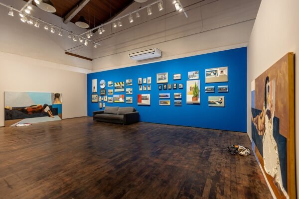 Vista da exposição de D.C. no Instituto Çarê. Sala ampla com piso de madeira escura. Uma parede azul exibe quadros de tamanhos variados. À esquerda e à direita, duas pinturas maiores retratam figuras desnudas sobre um sofá. Peças de roupa e calçados estão à frente das duas pinturas maiores, no chão.