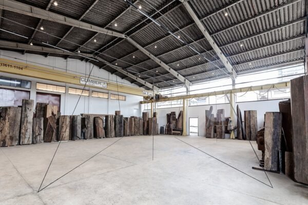 Vista da exposição de Fred Sandback no Instituto Çarê. Linhas finas em tecido preto atravessam o espaço do galpão com piso de concreto. Ao fundo, grandes troncos de madeira estão dispostos lado a lado.