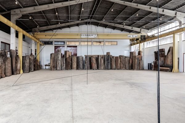 Vista da exposição de Fred Sandback no Instituto Çarê. Linhas finas em tecido preto atravessam o espaço do galpão com piso de concreto. Ao fundo, grandes troncos de madeira estão dispostos lado a lado.