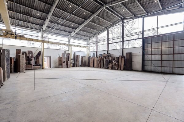 Vista da exposição de Fred Sandback no Instituto Çarê. Linhas finas em tecido preto atravessam o espaço do galpão com piso de concreto. Ao fundo, grandes troncos de madeira estão dispostos lado a lado.