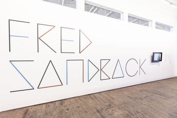 Vista da exposição de Fred Sandback no Instituto Çarê. A parede principal exibe o nome do artista, escrito com linhas minimalistas em formato geométrico, utilizando cores como preto, azul e laranja. Ao lado, há uma tela com vídeo exibido. O chão é de madeira, e o ambiente é bem iluminado.