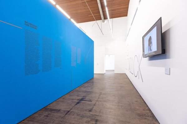 Vista da exposição de Fred Sandback no Instituto Çarê. Parede azul com texto explicativo à esquerda e monitor exibindo vídeo à direita. Espaço iluminado e piso de madeira.