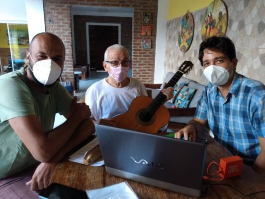 Yonan Gabriel, responsável pela transcrição musical, ao lado de Antonio Madureira e Francisco Andrade. Os três estão sentados em torno de uma mesa, com um violão em destaque, utilizado por Antônio. Francisco e Yonan utilizam máscaras faciais. O ambiente possui paredes de tijolos aparentes, quadros e almofadas coloridas.